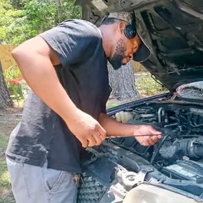 Person inspecting car engine with a tool in a wooded backyard, surrounded by trees and a wooden fence.