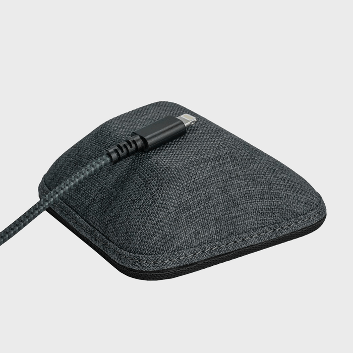 A compact speaker rests on a surface with a black cable connected, highlighting a modern design with textured fabric cover in neutral surroundings.