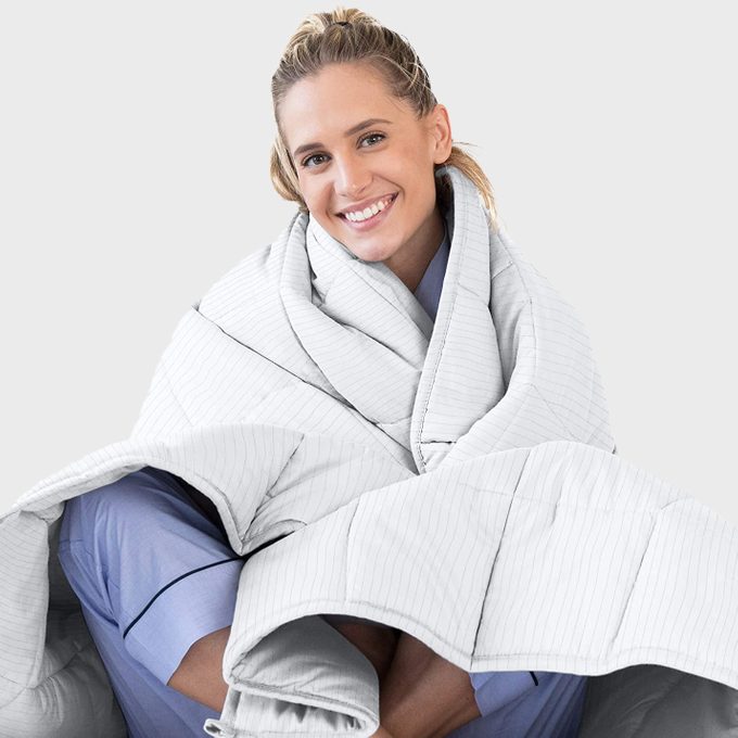 A person smiles while wrapped in a white striped blanket, wearing blue pajamas, against a plain grey background.