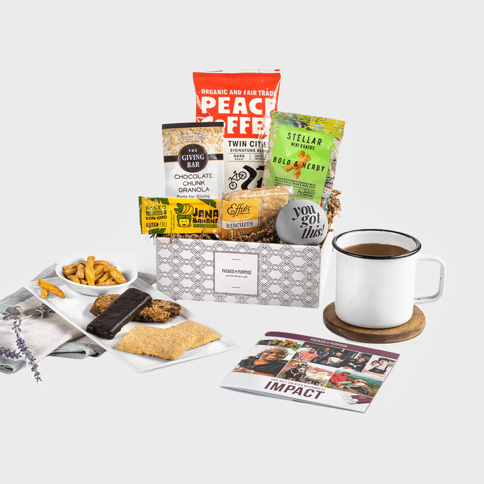 Gift box displays various snacks and coffee. Accompanied by a mug on a coaster and a pamphlet. Plain background enhances focus.