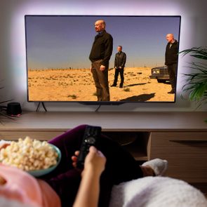 Person holding a remote and popcorn watches TV showing three men standing in a desert scene. Room features a small shelf and plants.