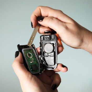 Hands holding a disassembled car key remote, revealing internal circuitry and battery, against a plain light blue background.