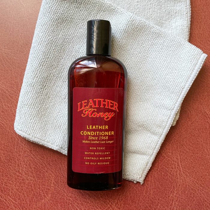 A bottle of Leather Honey conditioner rests on a white cloth on red leather. The text highlights its non-toxic, water-repellent, and mildew-controlling properties.