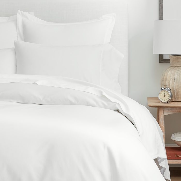A white bed neatly made with pillows, next to a bedside table holding a lamp and an alarm clock in a calm bedroom setting.