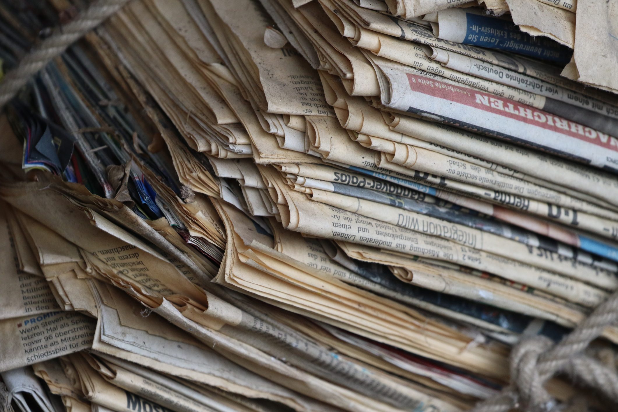 Stacked newspapers compressed by rope, aged and yellowing, in a tightly packed pile. Text is visible but not fully legible.