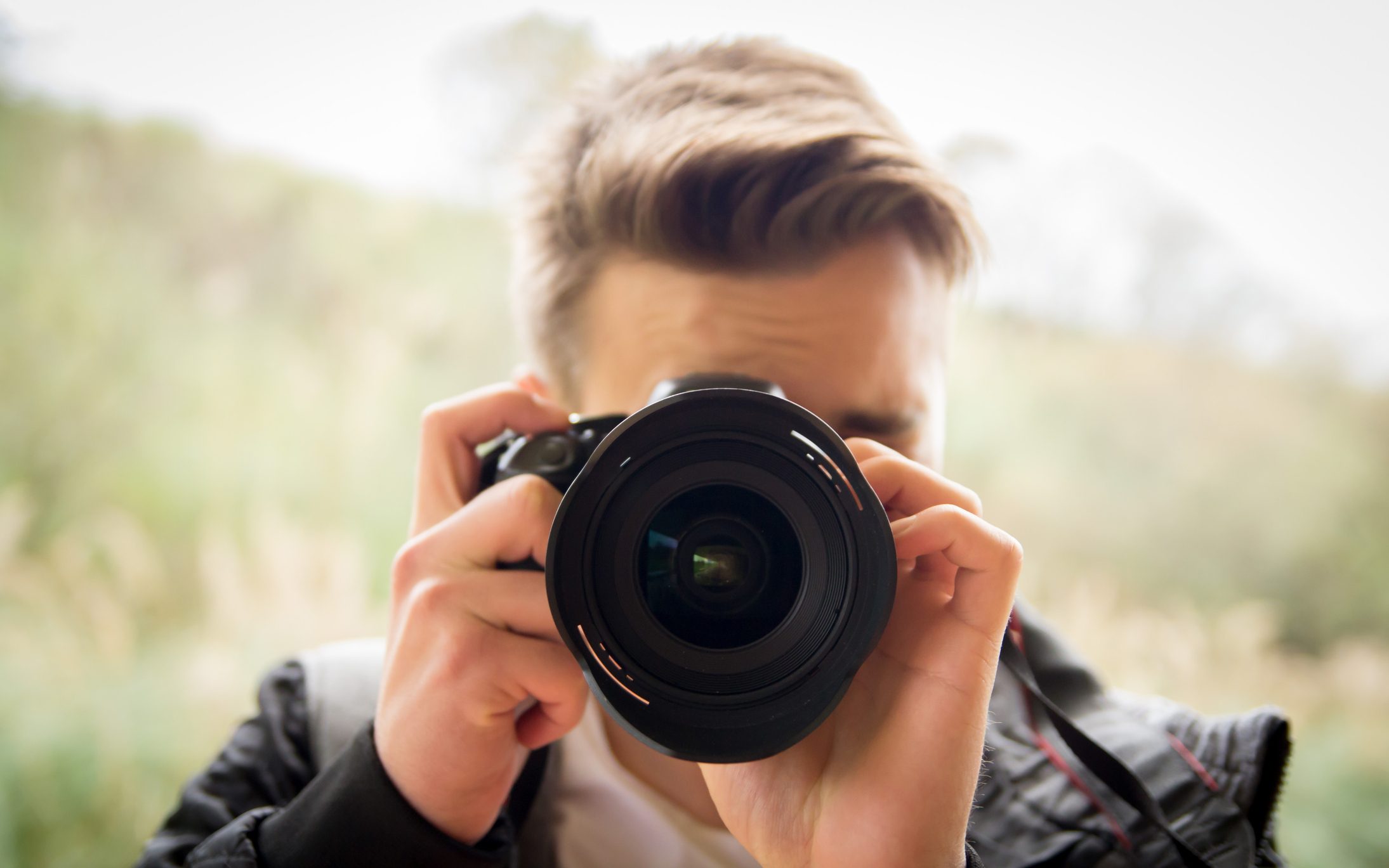Camera lens facing forward, held by a person, outdoors with a blurred natural background.