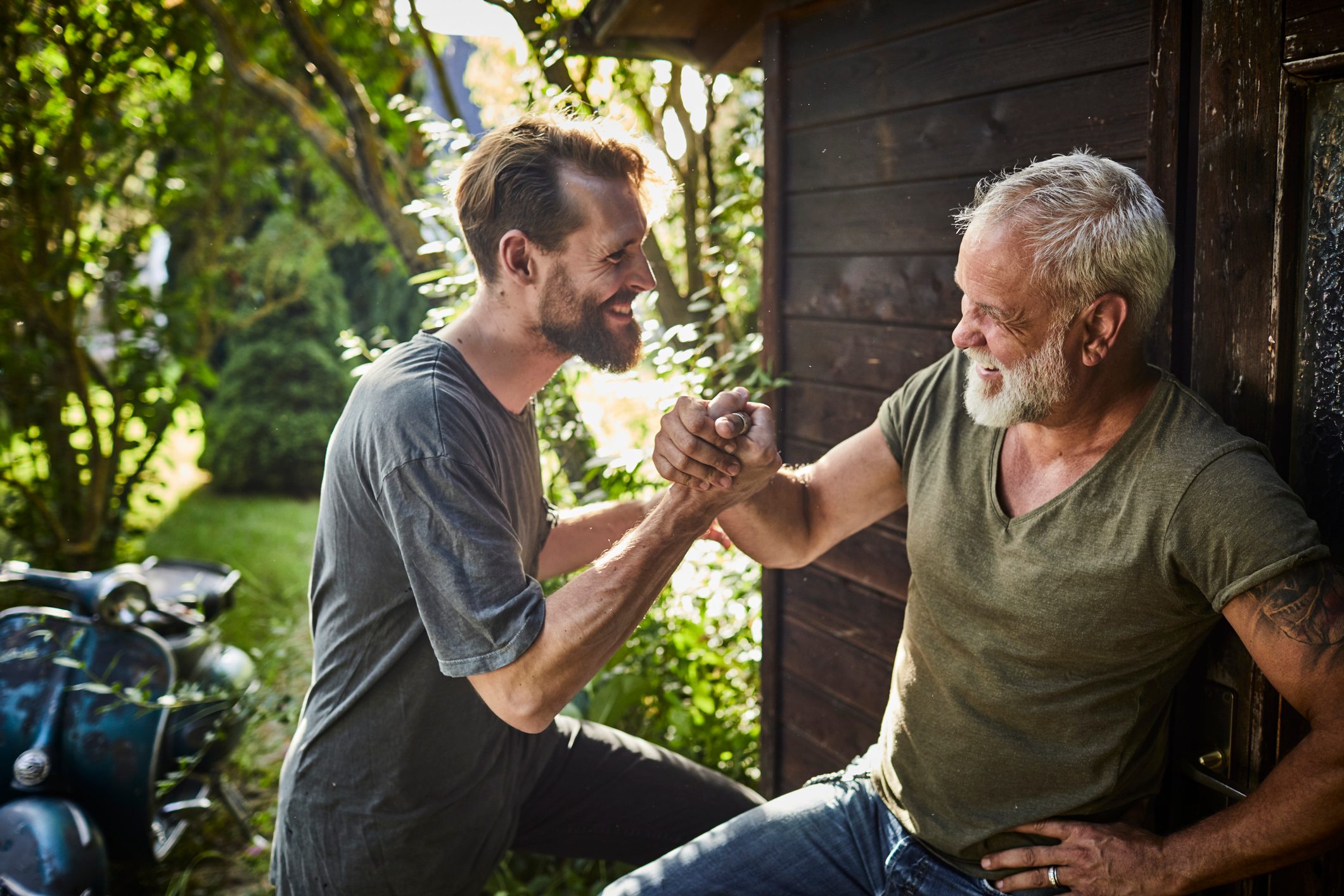 Two happy men shaking hands at garden shed with old motor scooter in background
