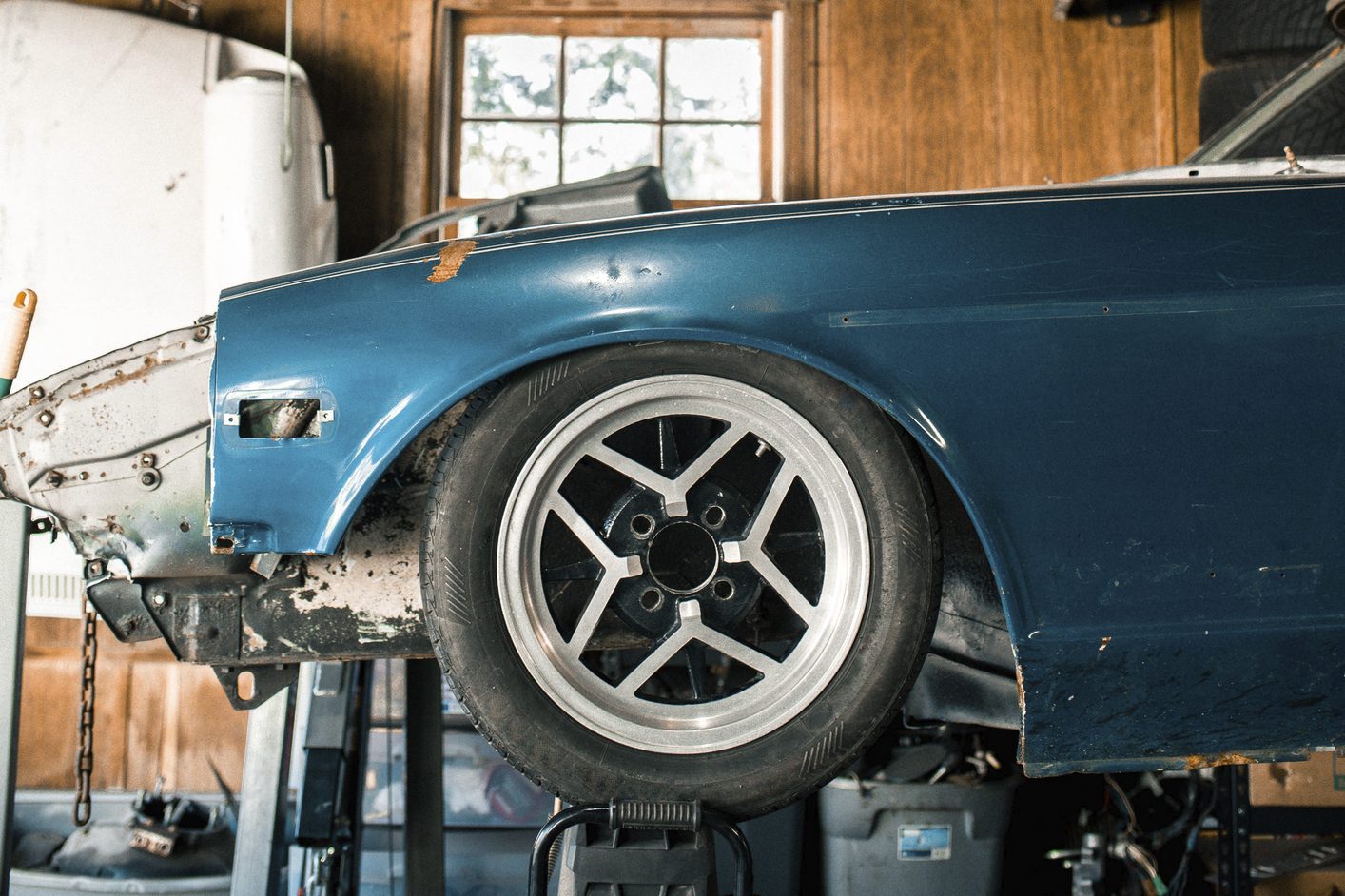 Car wheel rests on a lift, exposing a damaged blue fender in a wooden garage with a window.
