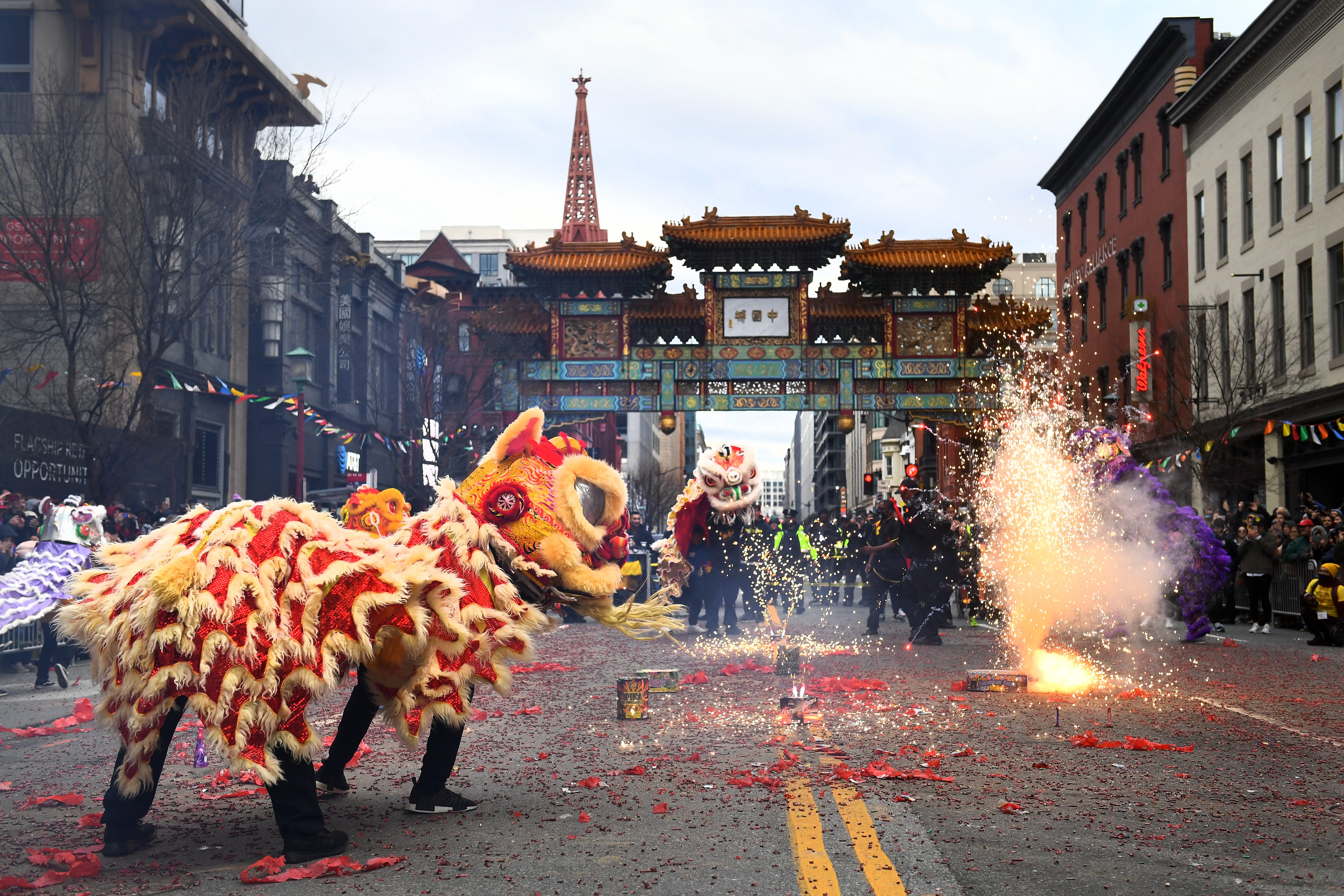 Dragon costumes dance, sparklers burst, amid a festive parade with colorful traditional archway and cheering crowd in a city street.