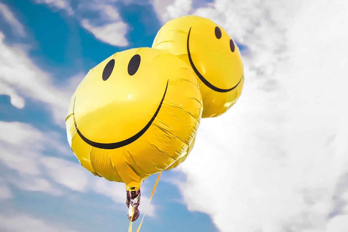 Yellow balloons float against a blue sky filled with scattered white clouds, each balloon displaying a large black smiley face.
