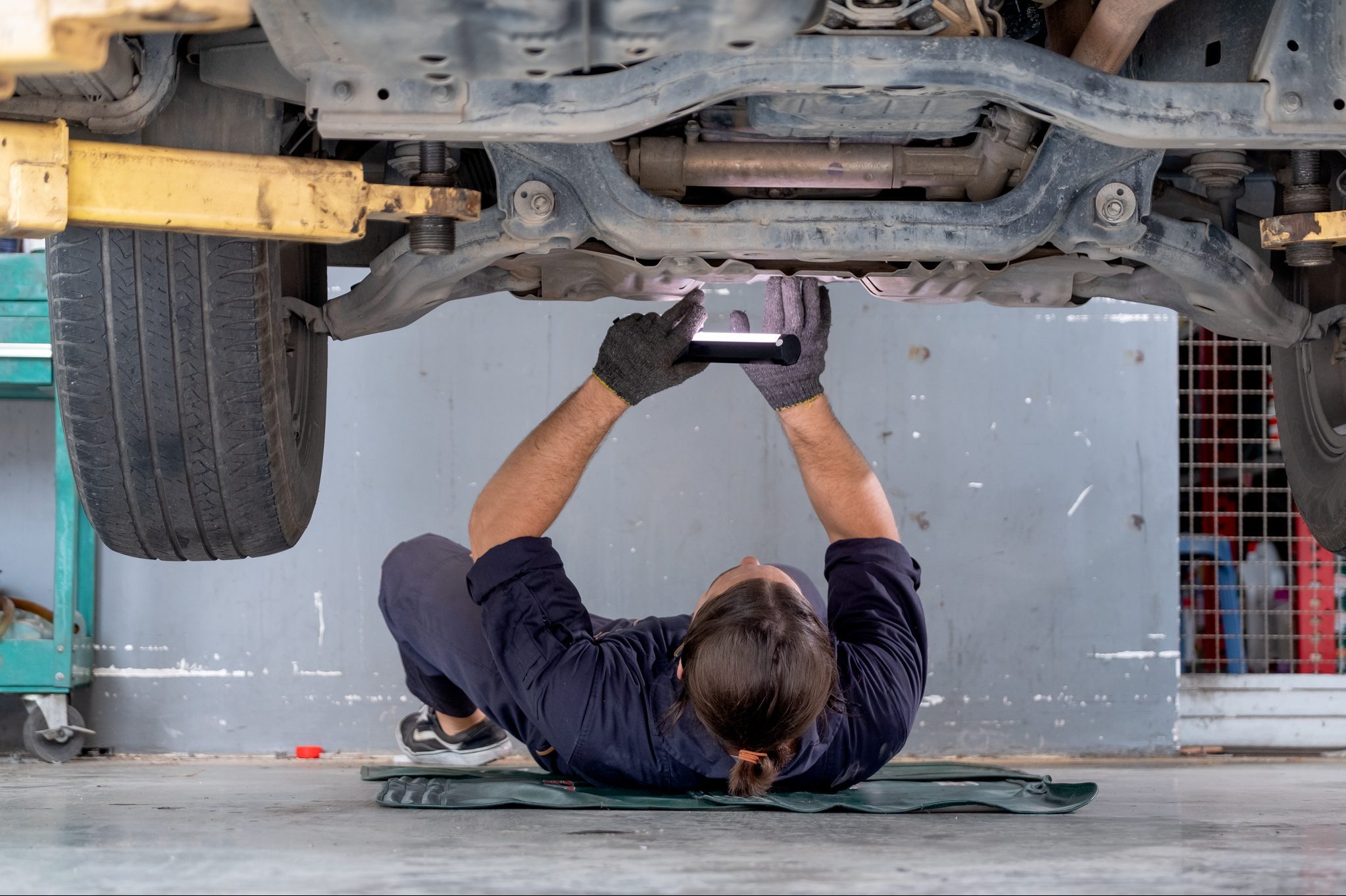 Mechanic examines vehicle undercarriage with flashlight in a garage, lying on a mat.