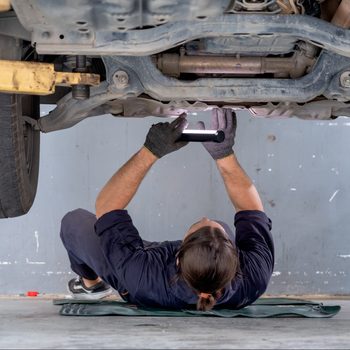 Mechanic examines vehicle undercarriage with flashlight in a garage, lying on a mat.