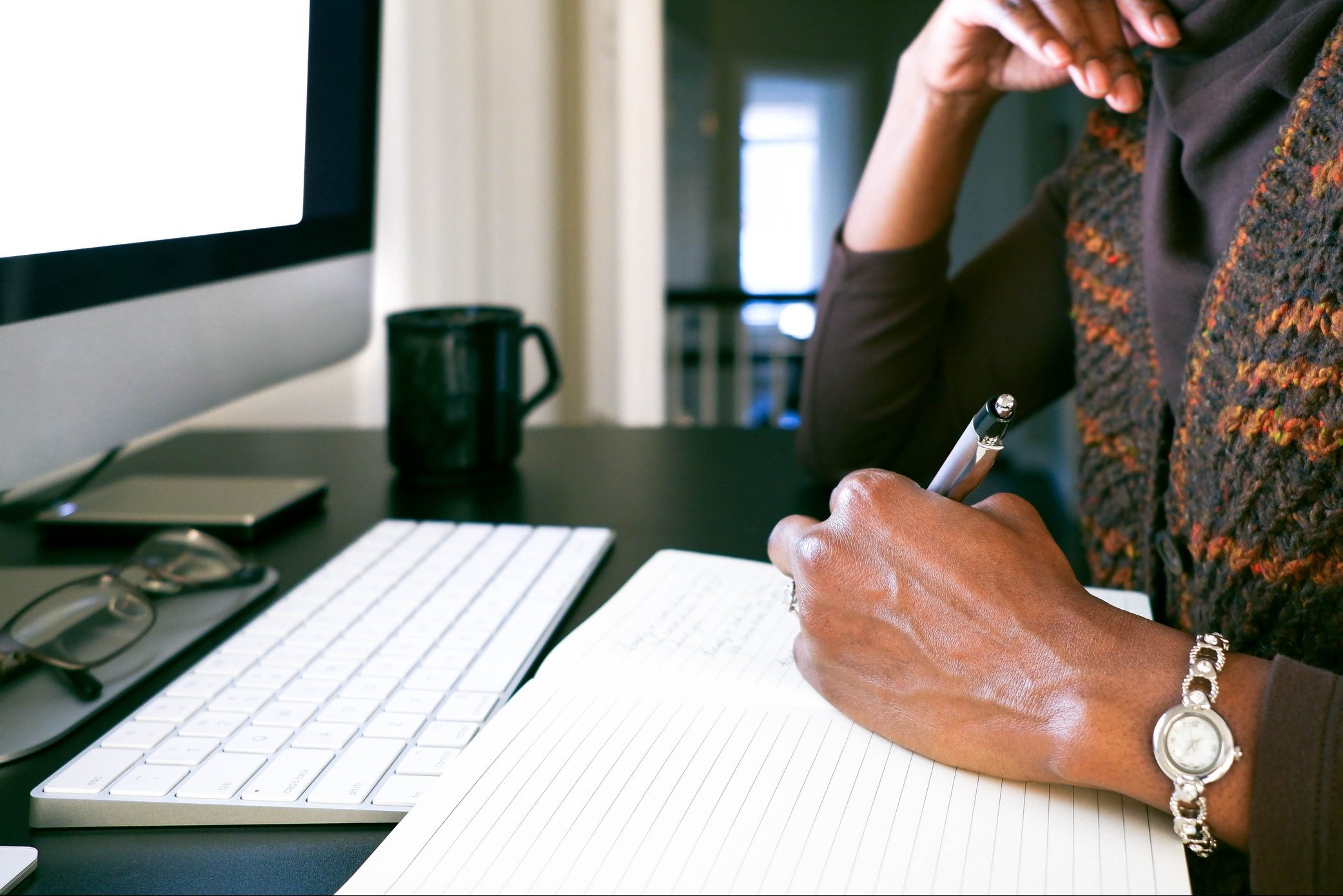 Person writing in a notebook beside a computer and keyboard, in an indoor workspace with a mug and glasses nearby.