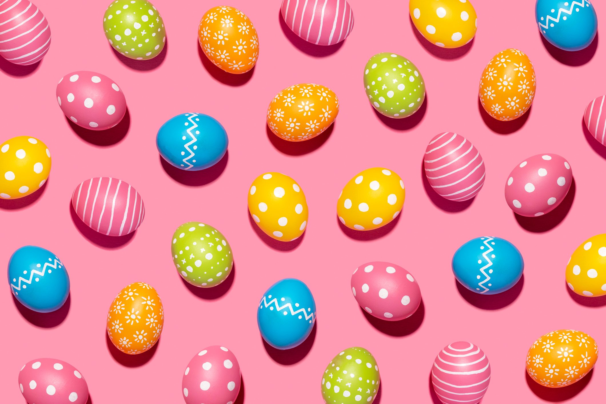 Colorful, patterned Easter eggs lie scattered on a pink background, showcasing various designs like stripes, polka dots, and zigzags.