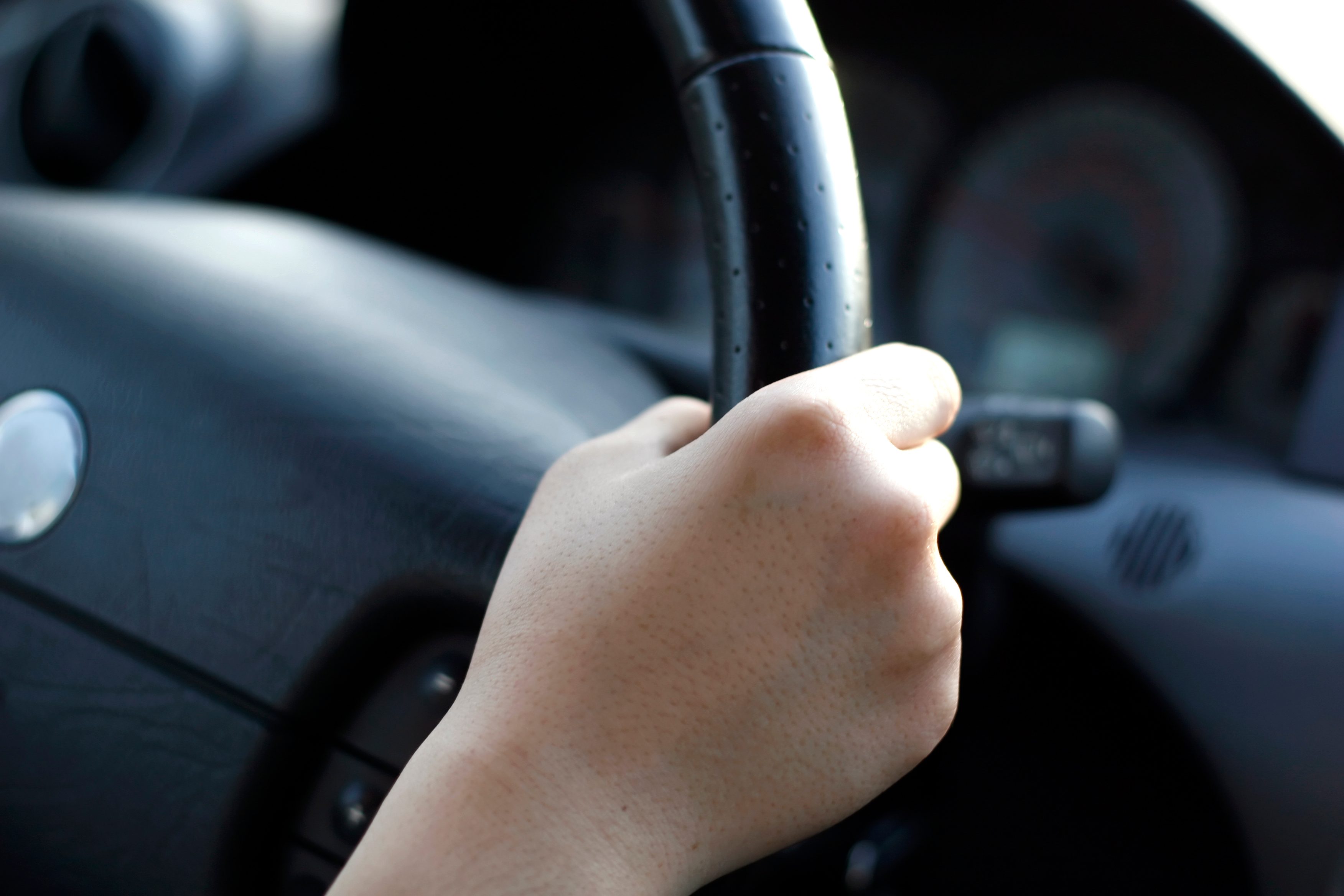 A hand grips a black steering wheel in a car interior; dashboard and air vent visible in the background.