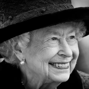 Elderly woman smiles warmly, wearing an ornate hat and pearl earring, against a blurred background.