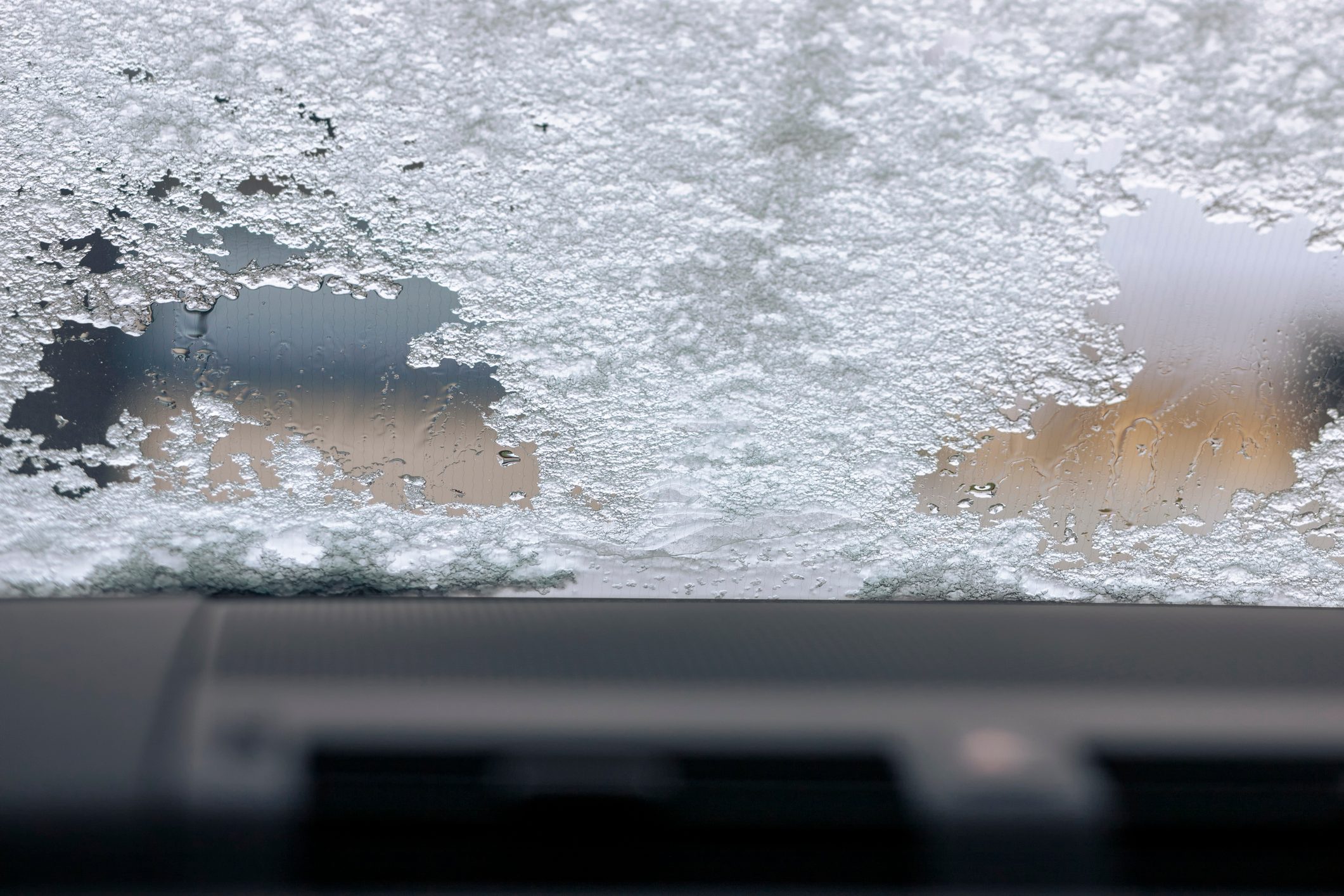 Snow covers a car windshield, partially melting, with the dashboard visible at the bottom edge.