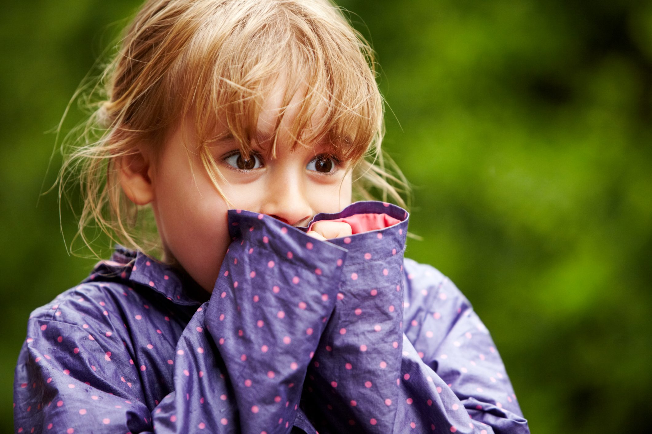 Child covers mouth with sleeve, eyes wide in surprise, outdoors, wearing a purple polka-dot jacket against a blurry green background.