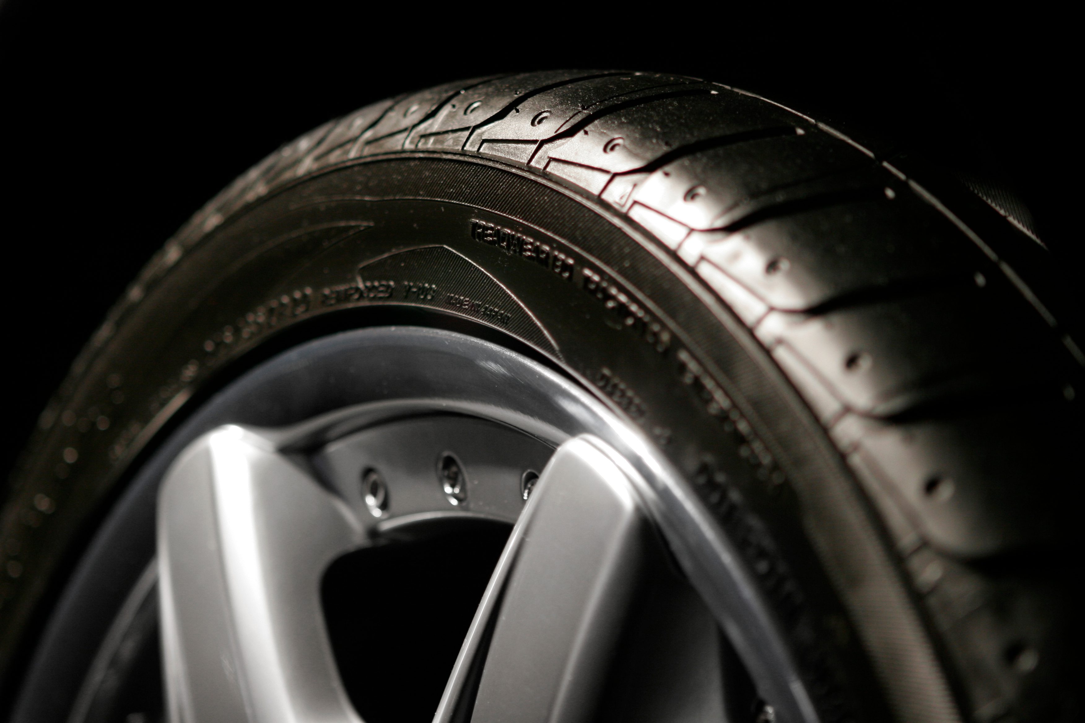 Tire sits still, illuminated by light, displaying detailed tread pattern, round metal rim visible, set against a dark background.