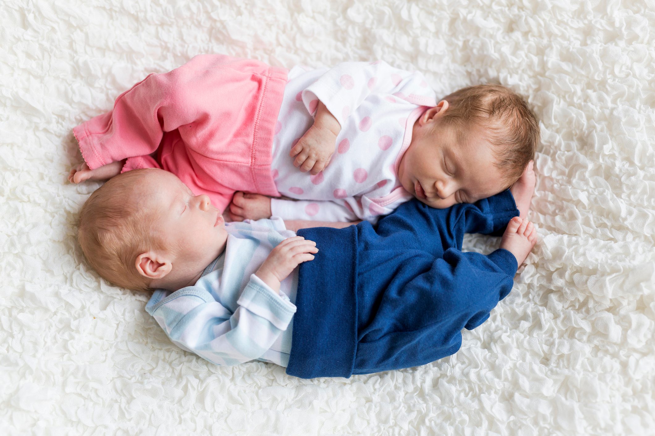 Two sleeping infants lie closely on a soft, white textured blanket, one wearing pink, the other blue, peacefully resting together.