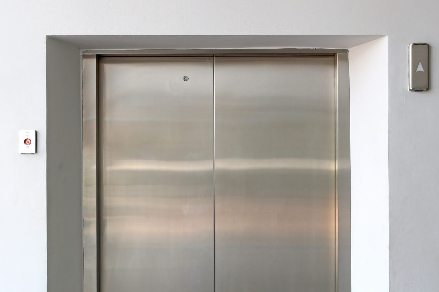 Elevator doors closed, in a white-walled hallway, with an upward arrow button on the right and a fire emergency button on the left.