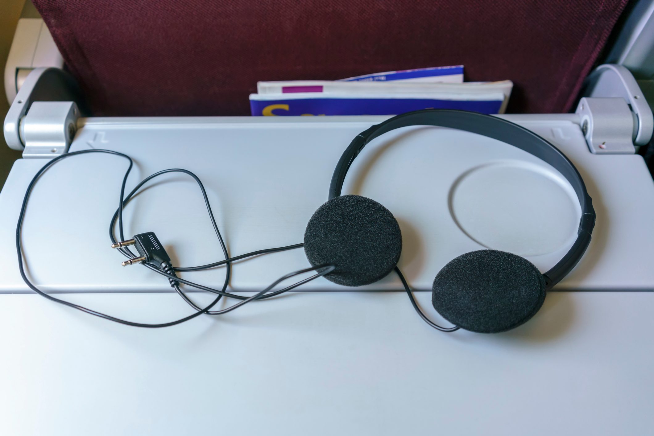 Headphones rest on a tray table, connected by wires. Airplane seat pocket behind holds magazines.