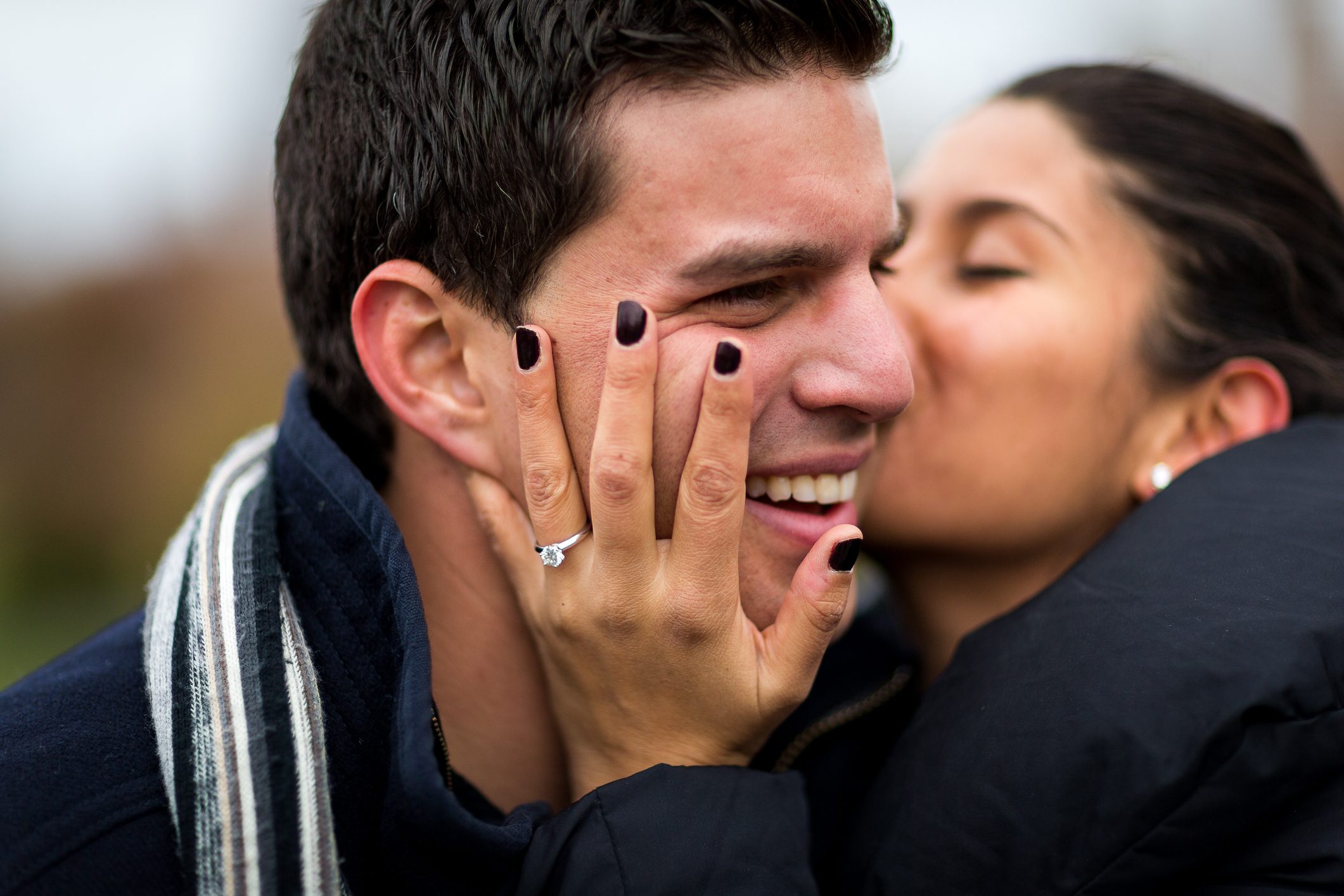 A woman kisses a smiling man, her hand with a ring on his cheek, outdoors in a close, affectionate embrace.