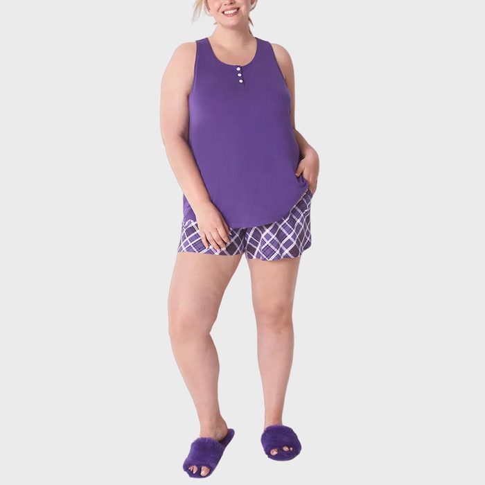 Woman wearing a purple tank top and plaid shorts, standing with one hand in pocket, against a plain background.