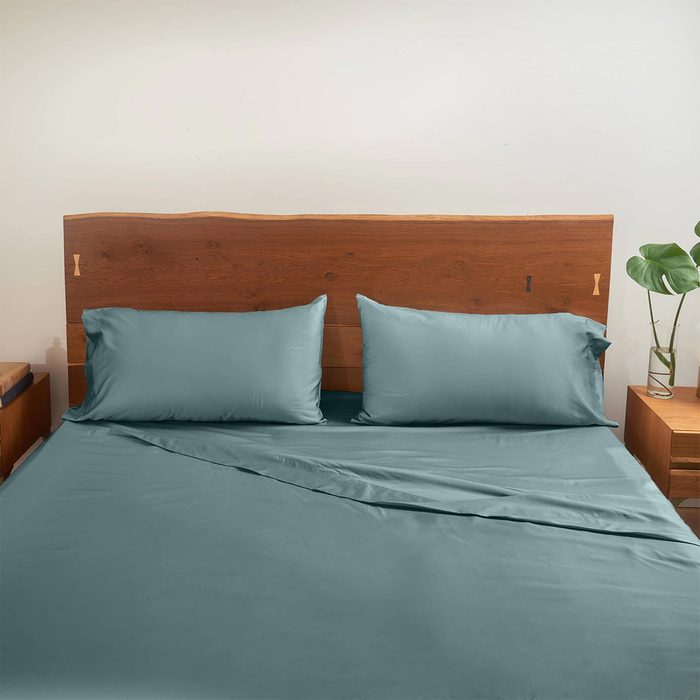 Bed displays teal sheets and pillows. The setting includes a wooden headboard and a bedside table with a small plant.