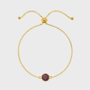 Golden chain bracelet with red gemstone centerpiece lies against a plain, light background. Two adjustable ends feature small gold beads.