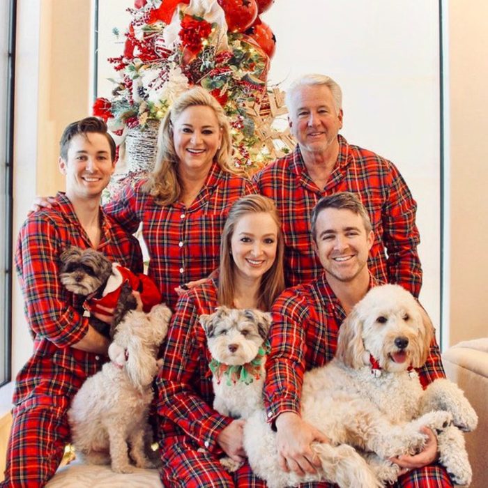 Family wearing matching plaid pajamas poses with dogs in front of a decorated Christmas tree indoors.