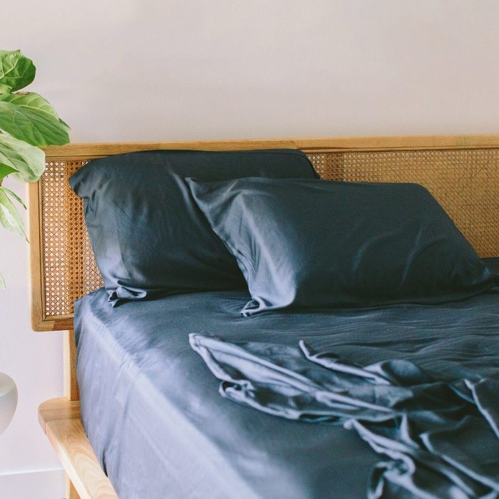 Bed with navy sheets and pillows, neatly arranged, in a room with a wooden headboard and a large green potted plant.