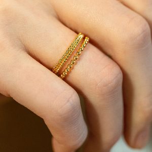 Two gold rings adorn a finger, one with a braided design and the other beaded, against a blurred background.
