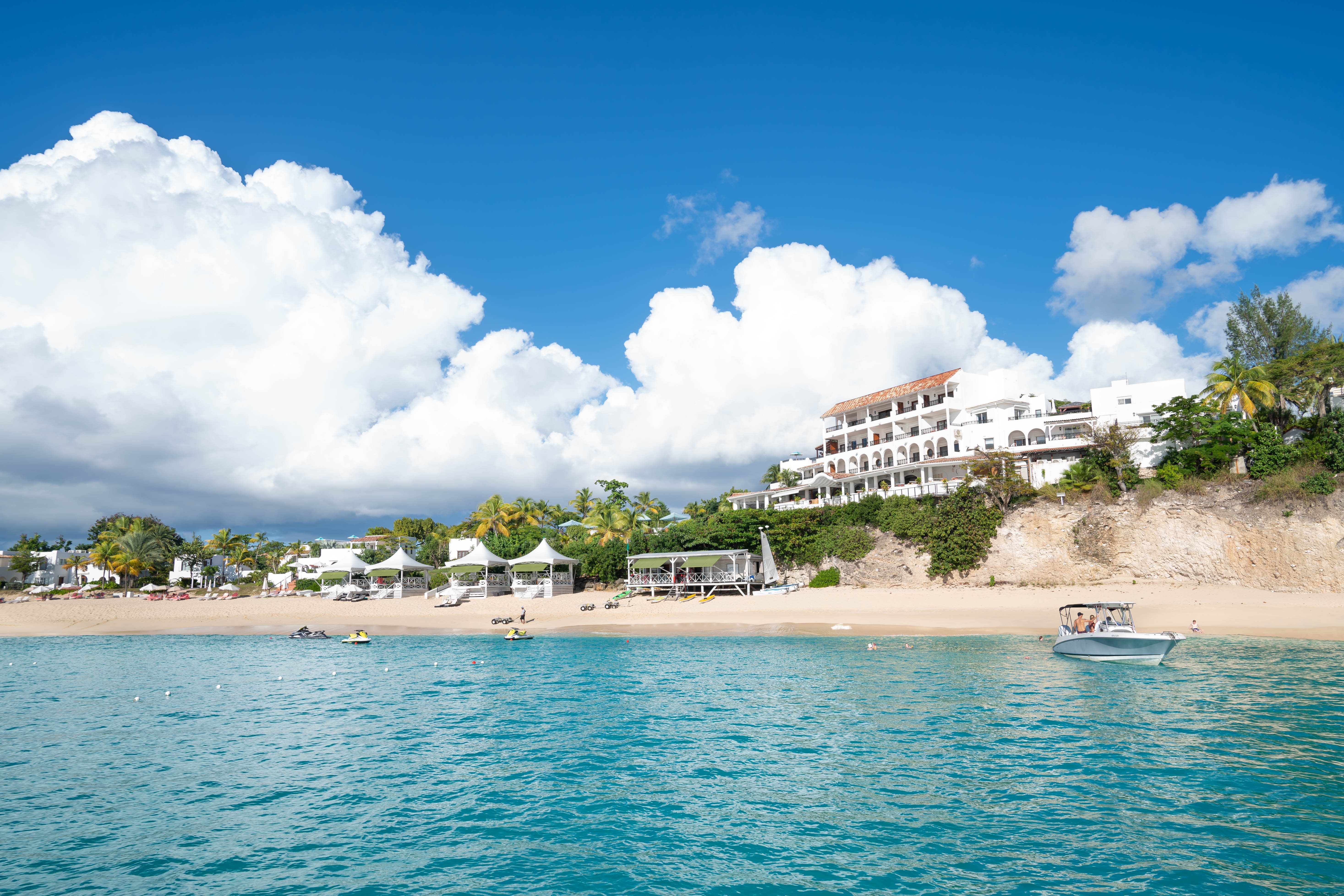 A boat floats on turquoise water near a sandy beach with white tents and a hillside resort, under a bright, partly cloudy sky.