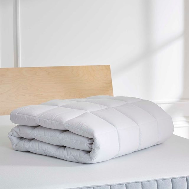 A folded, white quilt resting on a mattress in a minimalistic, well-lit room with a light wooden headboard.