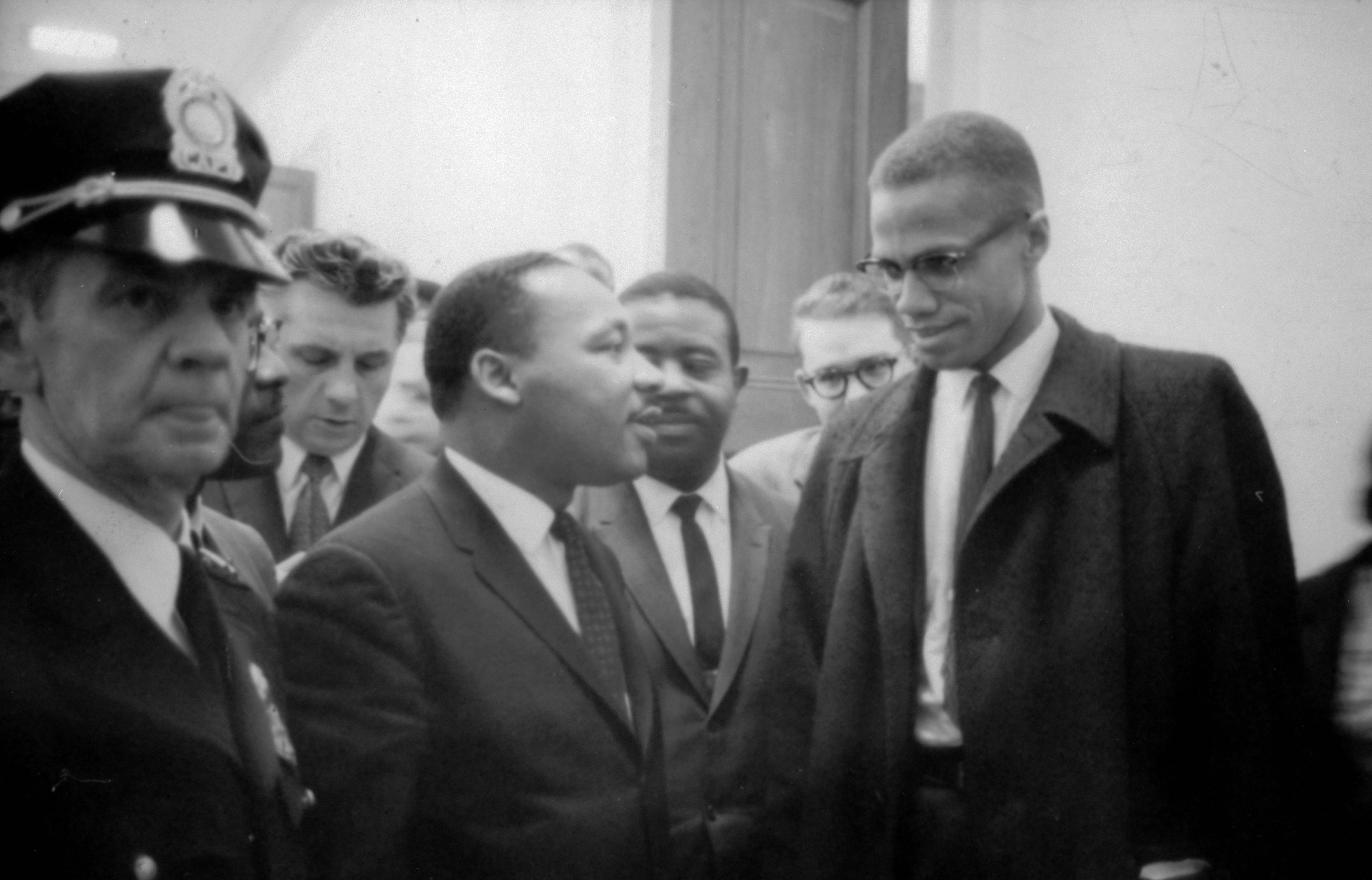 Two men in suits converse in a crowded hallway, surrounded by others, including a police officer in uniform.