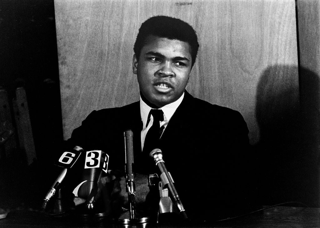 Man speaks at a press conference, wearing a suit, with microphones labeled "6" and "3" in front of a wooden background.