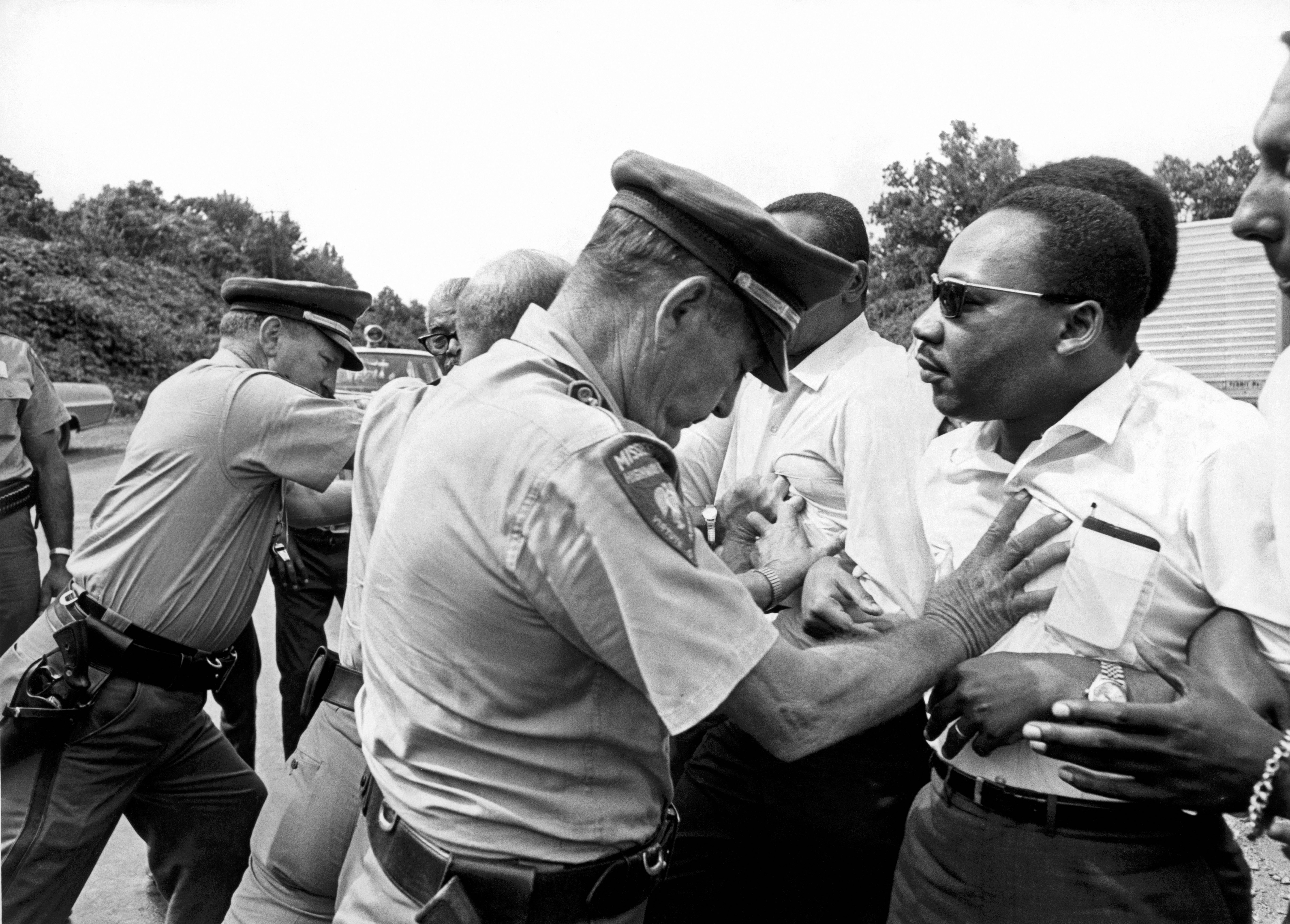 Officer restrains a man during a tense interaction. Several people are engaged, surrounded by trees and vehicles, in an outdoor setting.