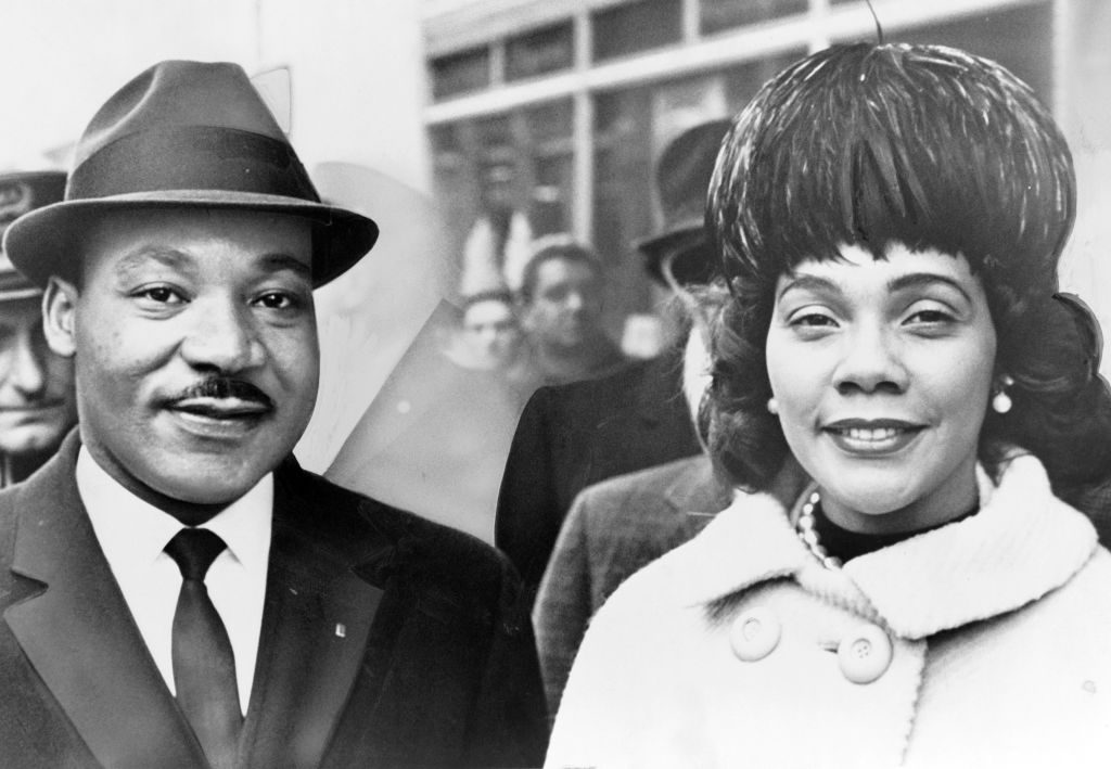 A man and woman smiling stand closely together; the woman wears a fur coat. They are outdoors with blurred figures and buildings behind them.