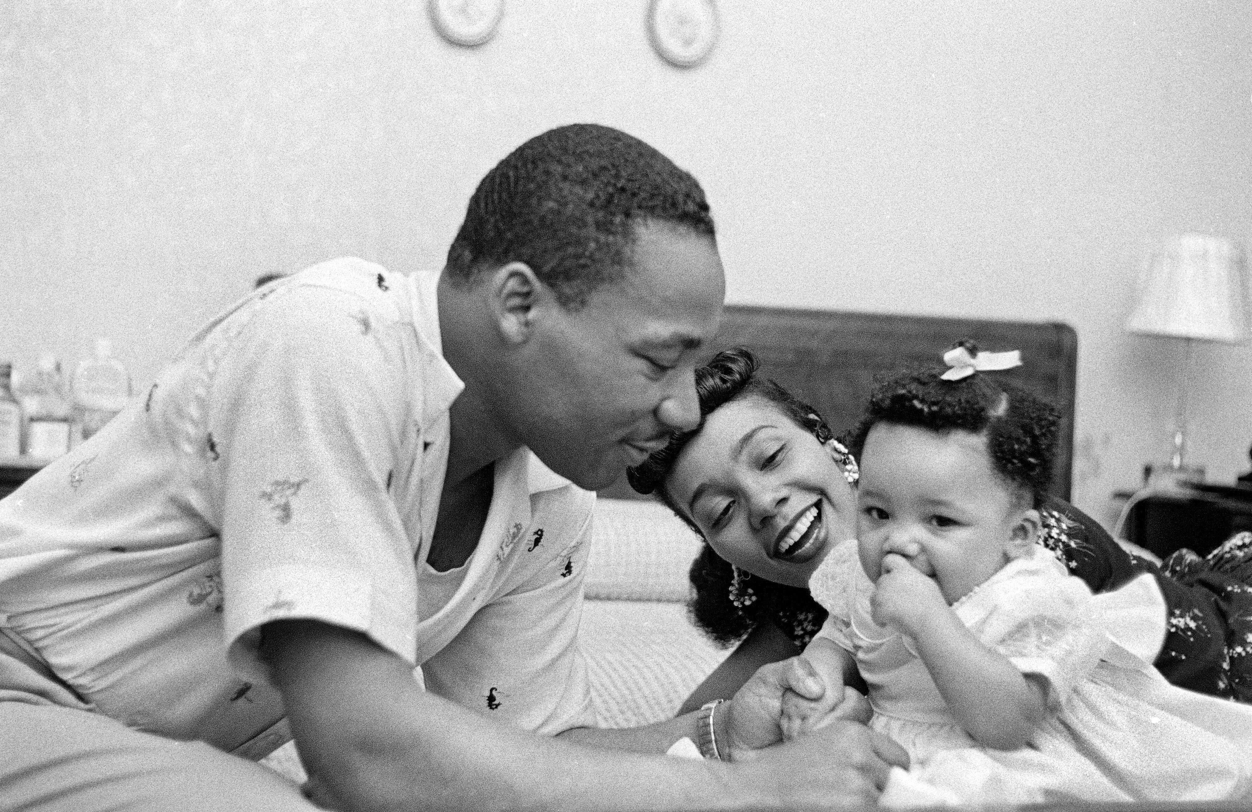 A man gently leans toward a woman and child on a bed, showing affection. A nightstand with a lamp is in the background.