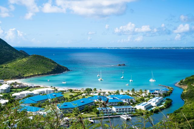 Sailboats float calmly in a turquoise bay, surrounded by lush green hills and blue-roofed buildings under a clear sky.