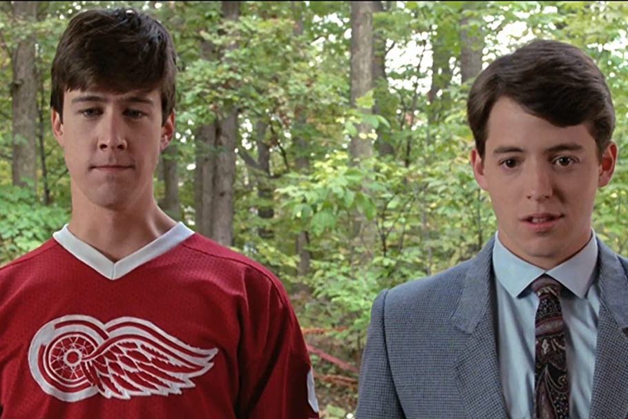 Two young men stand side by side, one wearing a red sports jersey. They are outdoors, surrounded by greenery and trees.
