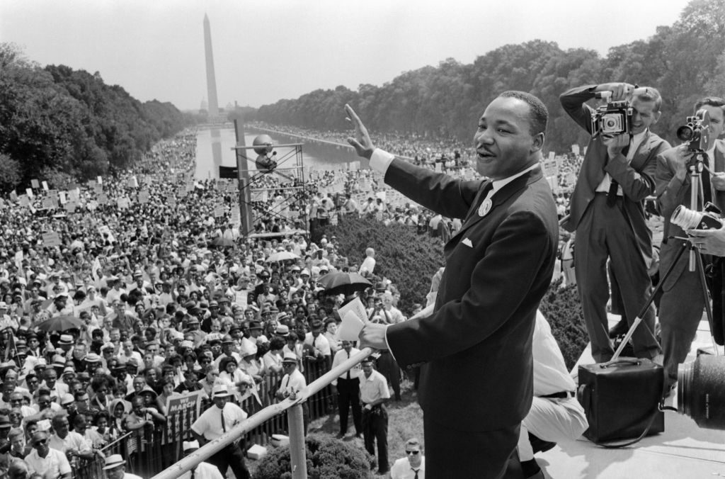 A man speaks passionately to a large crowd gathered around the reflecting pool in Washington, D.C., with photographers capturing the event.