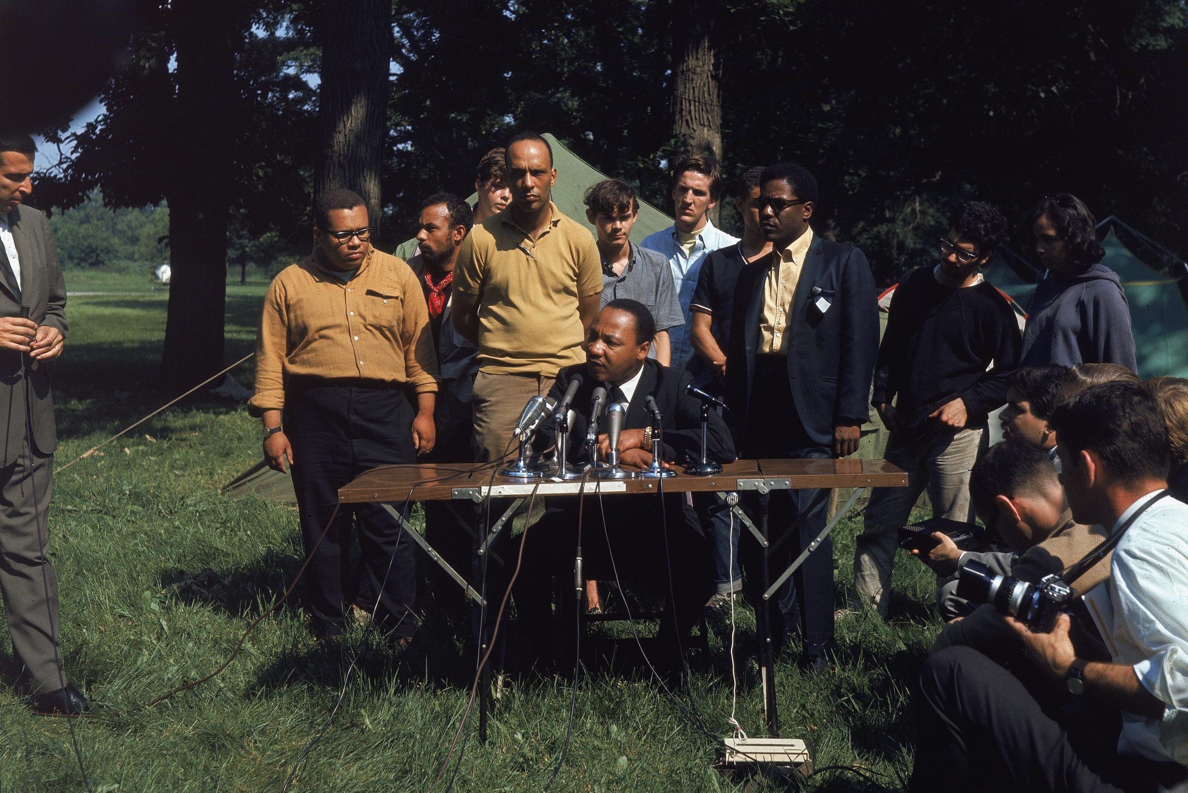 Man speaks at outdoor press conference; surrounded by people and reporters with cameras in a grassy setting. Several microphones are positioned on the table.