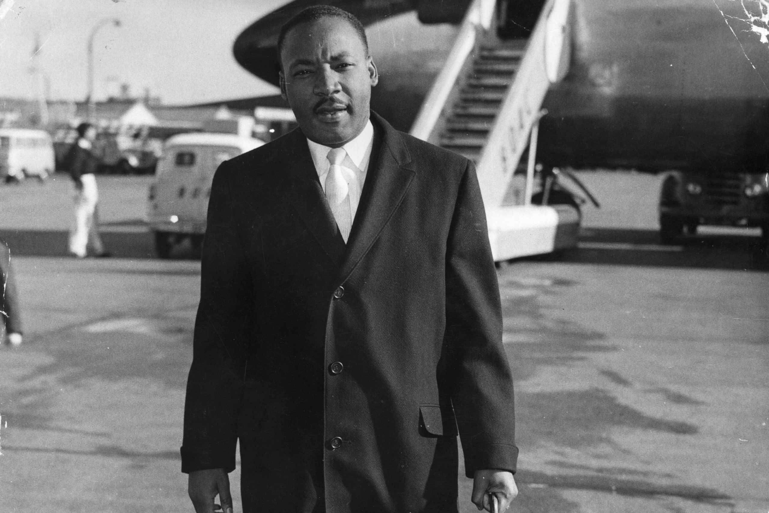 A man in a suit stands near an airplane's stairs at an airport, with vehicles and people in the background.