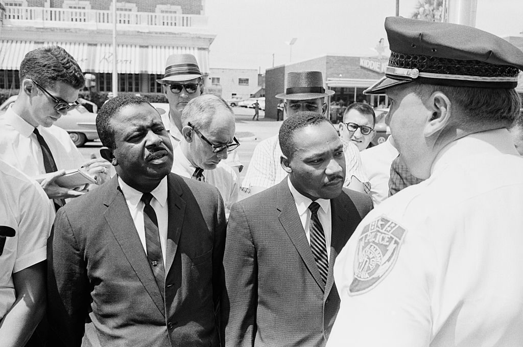 Two men, wearing suits, converse with a police officer outside; surrounded by onlookers and reporters in a city street.