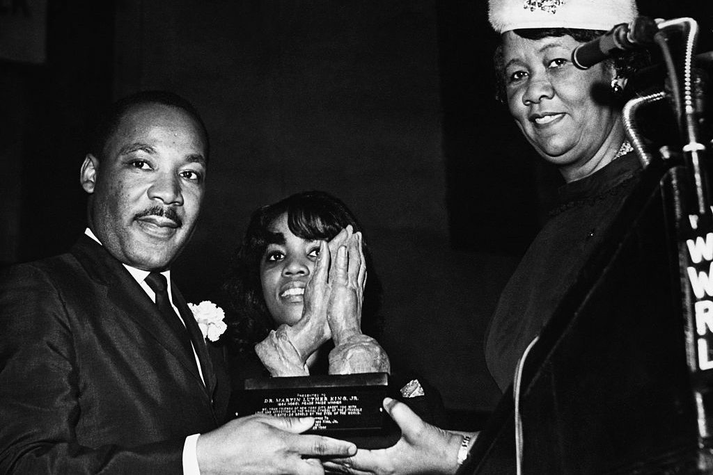 A man and two women hold a plaque together at a formal event, smiling, with a microphone on the right.