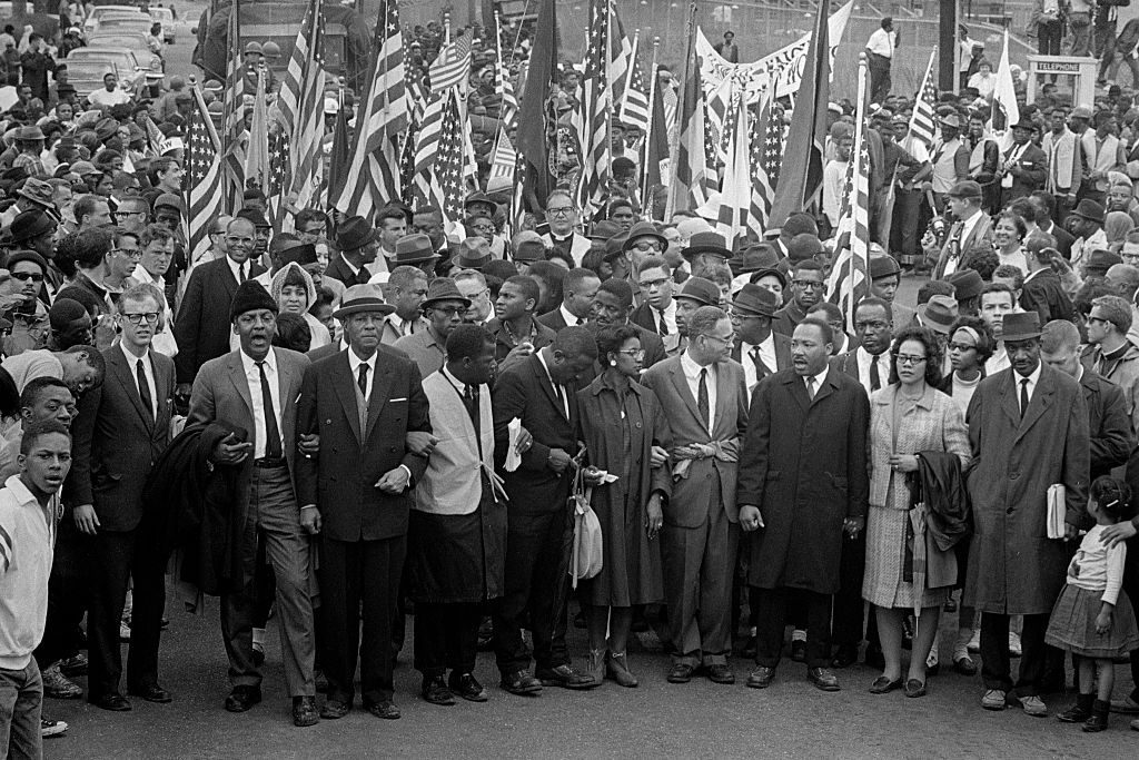 A diverse group marches with linked arms, holding flags, surrounded by a crowd in an outdoor setting.