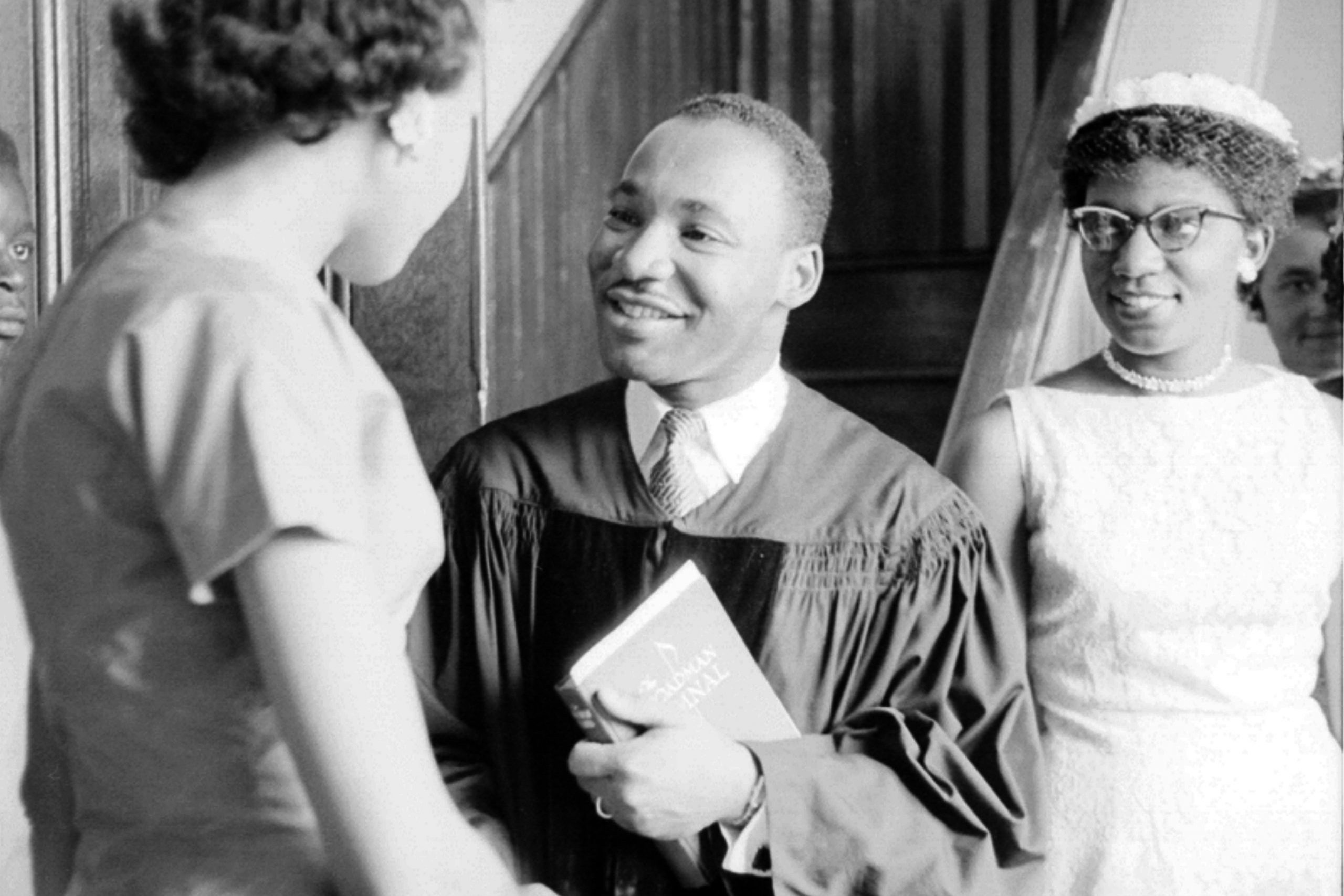 A man in a robe smiling and holding a book talks to a woman indoors, with another woman nearby.