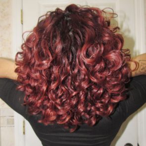 Curly red hair flows down a person's back, with arms raised, in front of a white door.